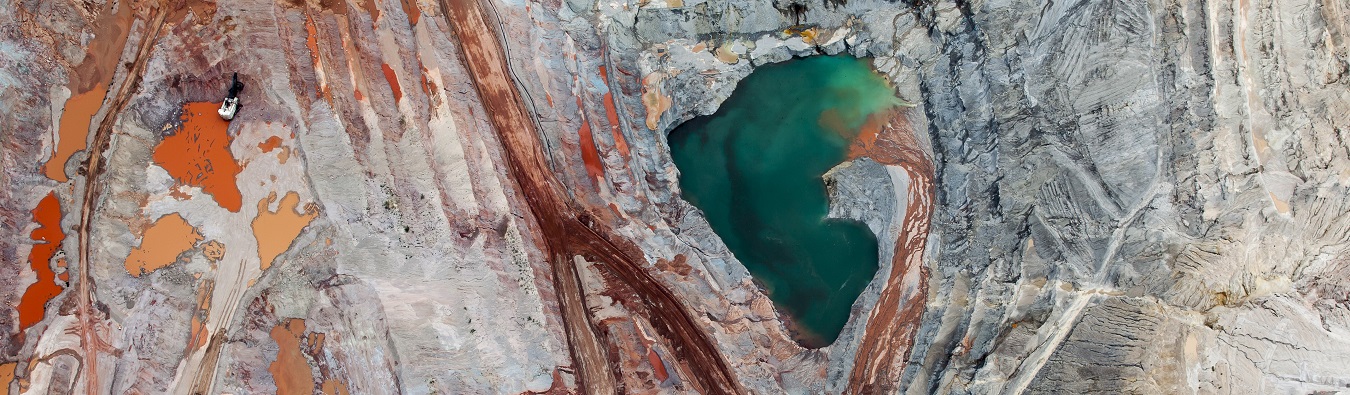 Aerial view of a mining site with distinct layers of red, orange, and gray earth surrounding a vibrant green water pool. The image showcases the vast scale and intricate patterns of the mining landscape, representing the transformative impact of infrastructure projects like the Simandou mining venture in Africa.